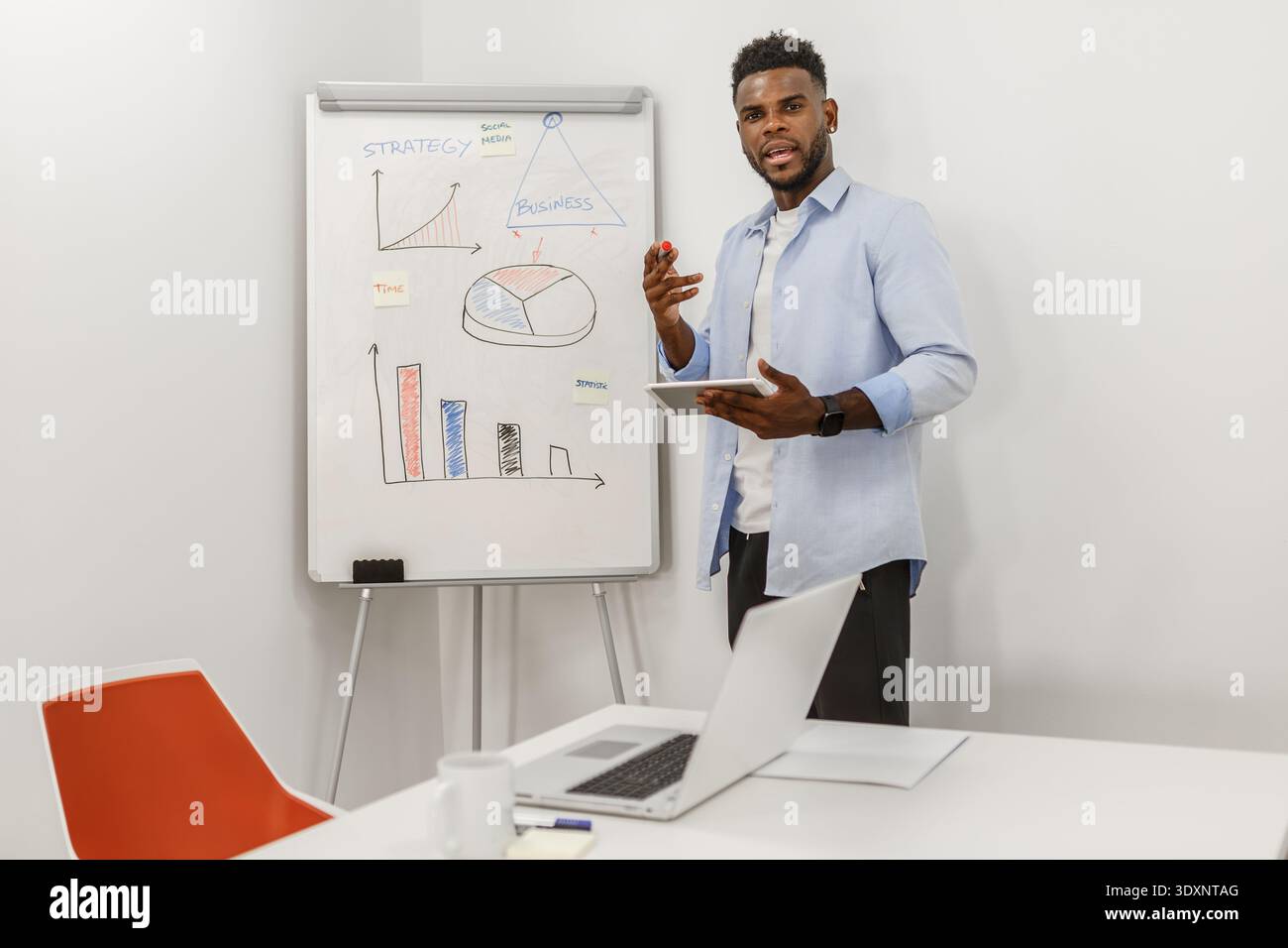 Ein Mann präsentiert eine Geschäftsstrategie in einem hellen Büro, verwendet ein Tablet und veranschaulicht Konzepte auf einem Whiteboard mit Diagrammen und Diagrammen. Hallo Stockfoto