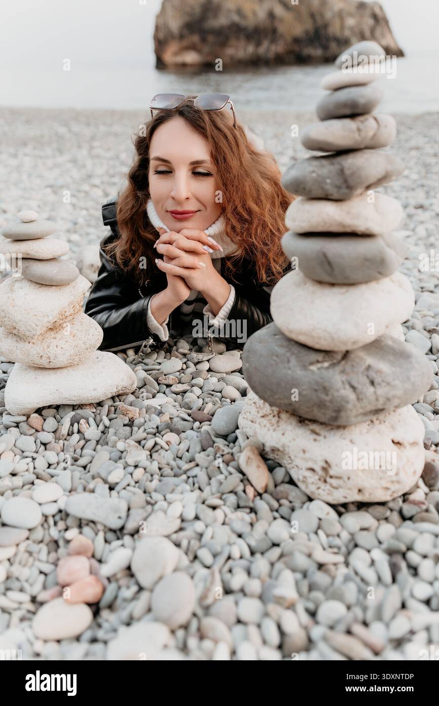 Frau mit Steinkieseln entspannt rothaarig liegend an einem felsigen Ufer mit gestapelten Steinen für eine meditative Zen-Atmosphäre Stockfoto