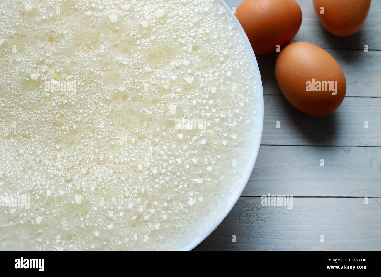 Eine weiße Schüssel mit geschlagenem Eiweiß, mit einer schaumigen Konsistenz und zahlreichen Luftblasen, auf grauem Holzhintergrund. Kulinarisches Kreativkonzept. Stockfoto