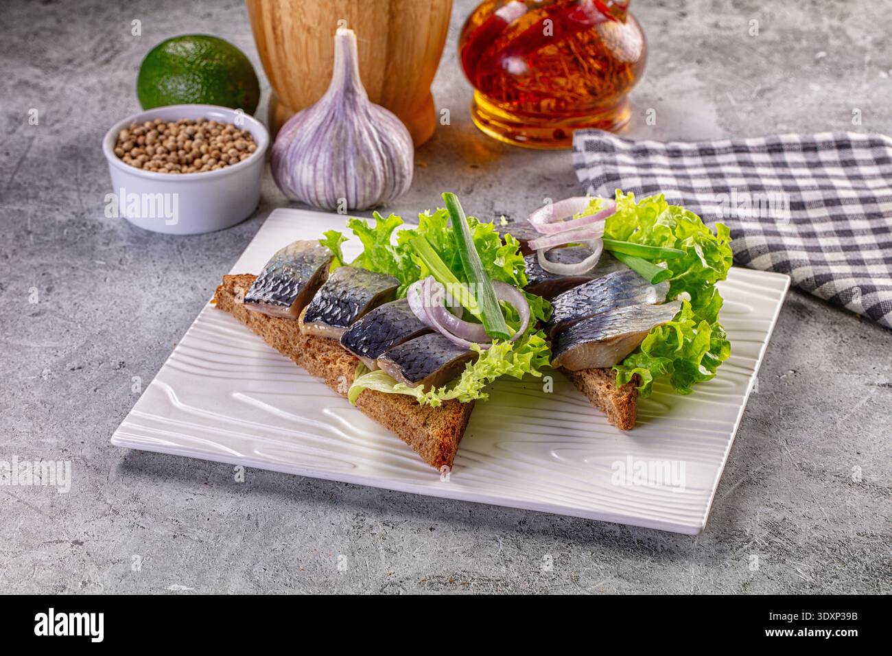 Vorspeise mit eingelegtem Hering auf dunklem Roggenbrot mit Salat und Zwiebeln. Serviert auf einem weißen Teller auf grauem Hintergrund mit Knoblauch und Gewürzen. Rustikales Foo Stockfoto
