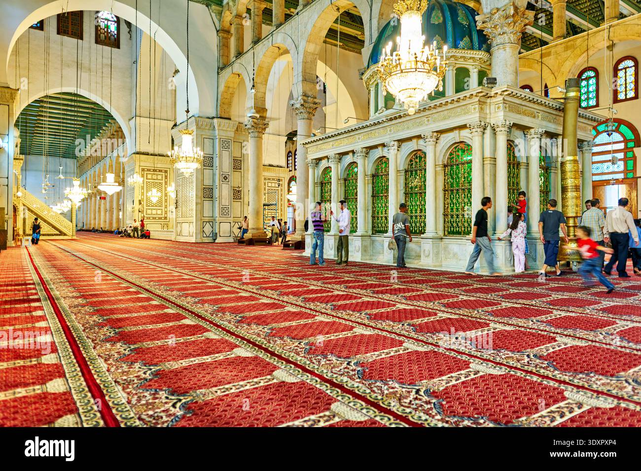 Die Menschen laufen durch das geräumige Innere der Umayyaden-Moschee in Damaskus. Die Gegend hat wunderschöne Bögen und einen reichen Teppich. Stockfoto