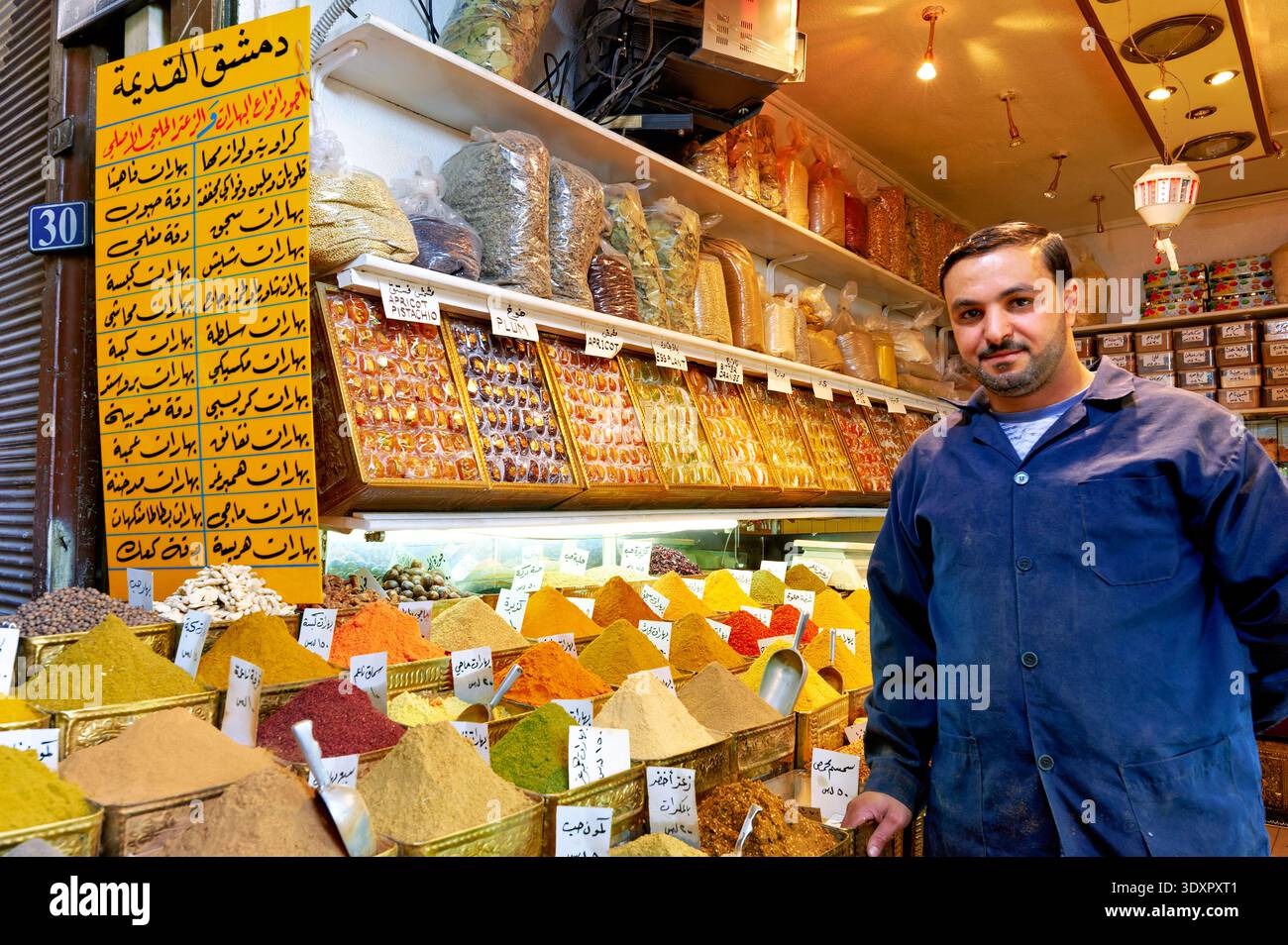 In Al Silah Souq steht ein Händler in der Nähe einer Ausstellung verschiedener Gewürze in Damaskus. Kunden stöbern durch die bunte Auswahl an Gewürzen. Stockfoto