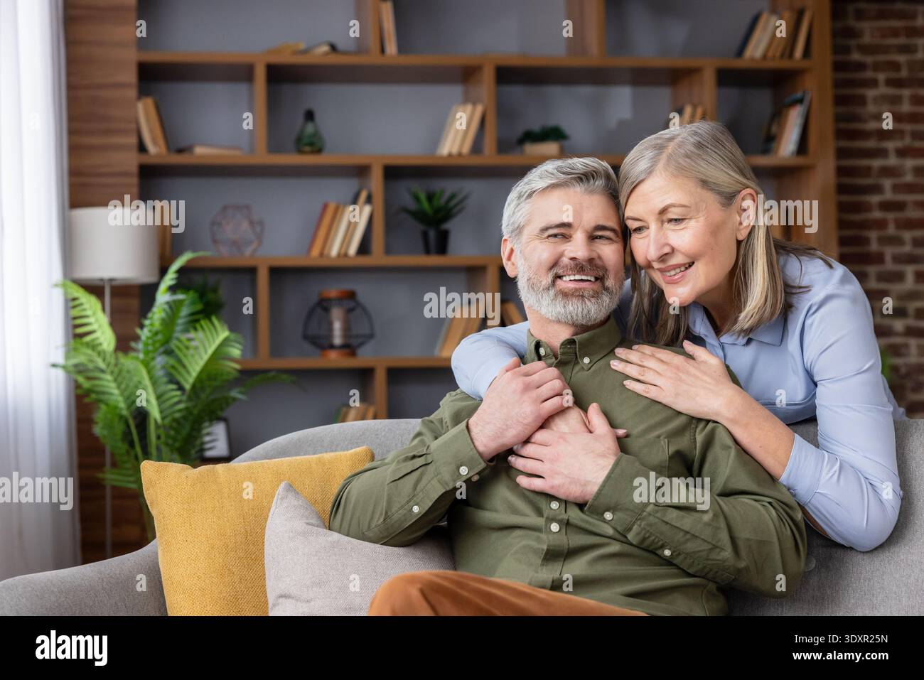 Ältere Paare sitzen auf einem Sofa in ihrem Wohnzimmer, umarmen und lächeln und repräsentieren Konzepte von Liebe, Partnerschaft, Glück und einem komfortablen Lebensstil Stockfoto