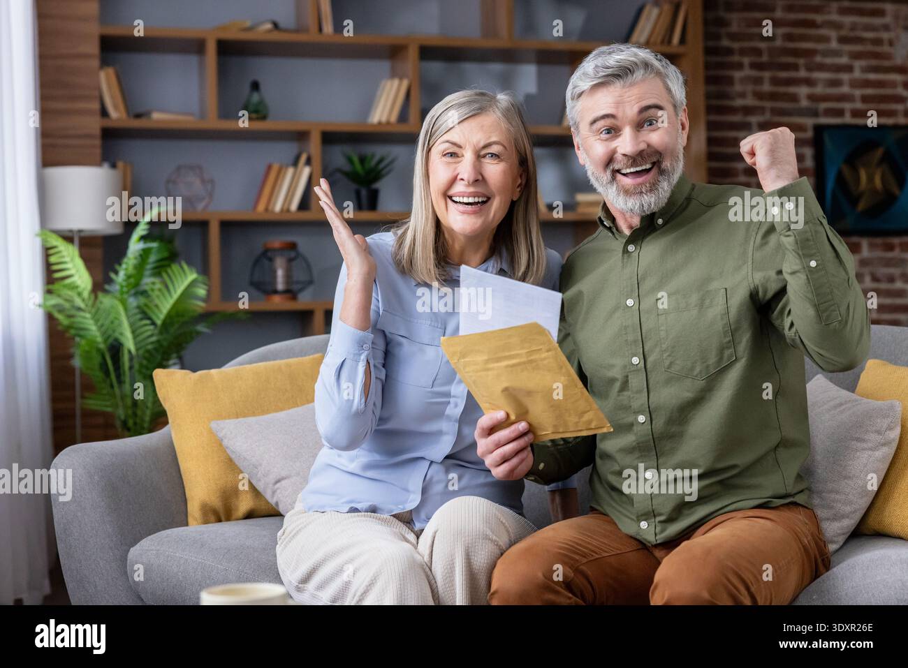 Ein aufgeregtes Seniorenpaar sitzt auf einem Sofa, liest wichtige Post und jubelt mit fröhlichen Ausdrücken, feiert finanziellen Erfolg, eine Investitionsrendite oder eine Ruhestandsankündigung zu Hause Stockfoto
