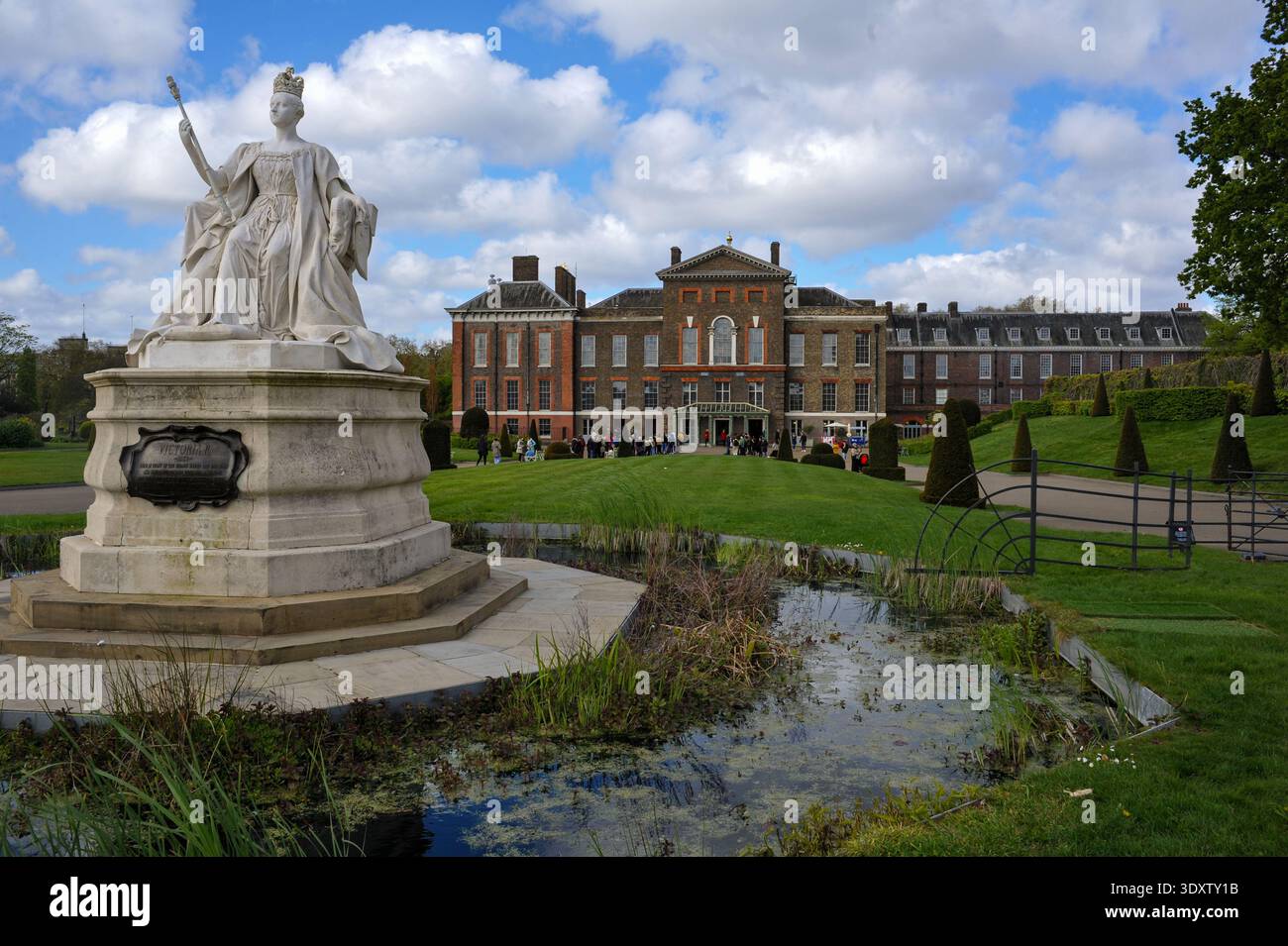 Statue der Königin Victoria auf einem Steinsockel vor dem Kensington Palace, mit Teich und Besuchern im Hintergrund an einem teilweise bewölkten Tag. Stockfoto