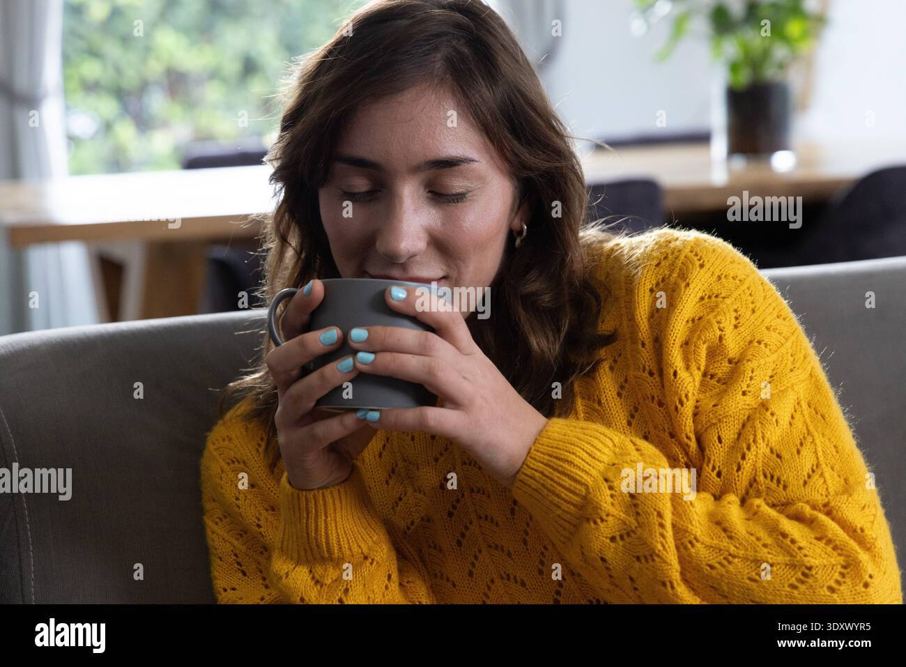 Frau in ihren 20ern in senfgelbem Strickpullover mit grauem Becher, das Aroma im Wohnzimmer einatmet Stockfoto