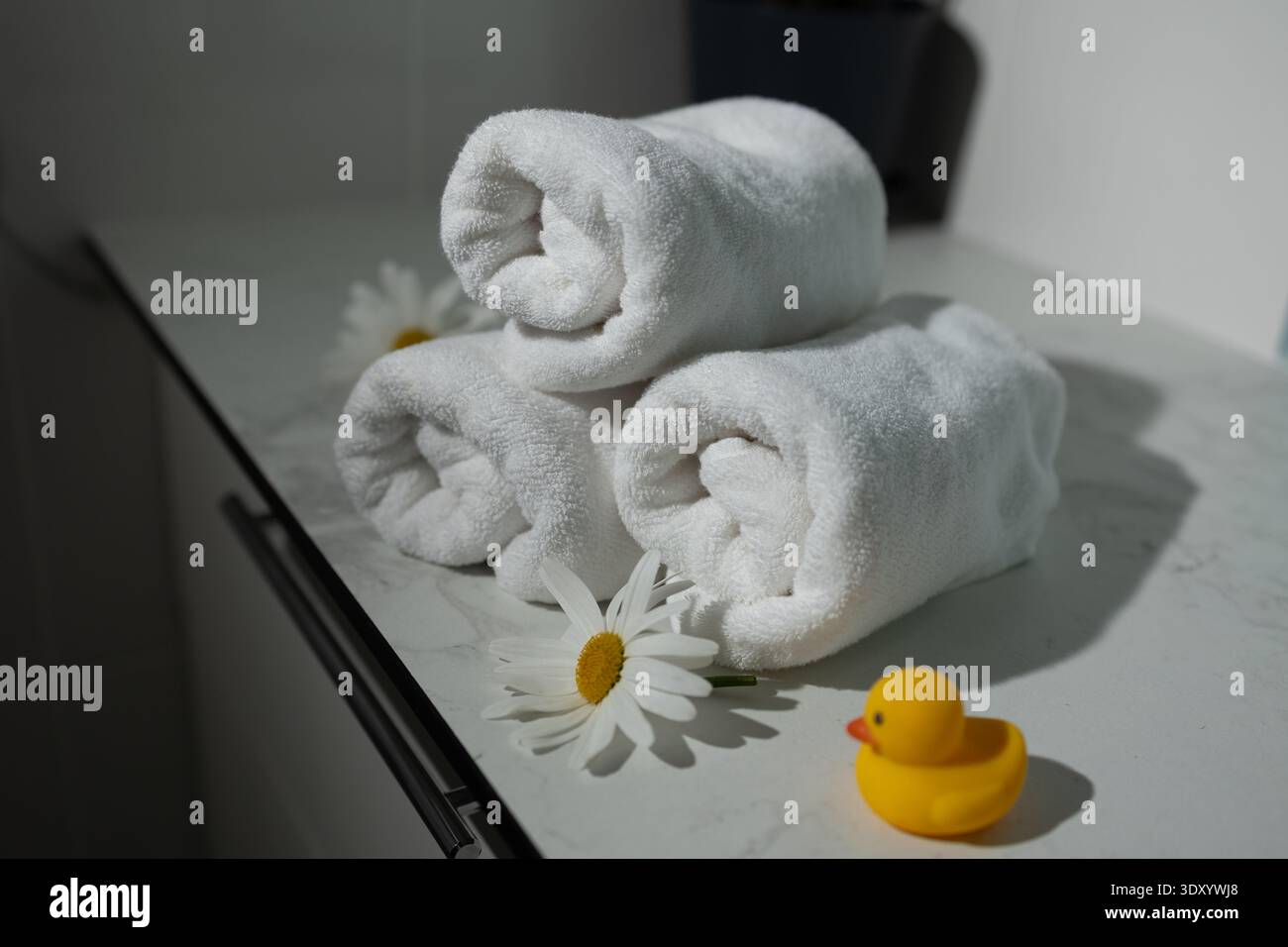 Weiche weiße Handtücher sind in einem ordentlichen Stapel neben zwei Gänseblümchen und einer verspielten Gummiente angeordnet. Dieses ruhige Spa-Ambiente lädt zu Entspannung und Komfort ein. Stockfoto