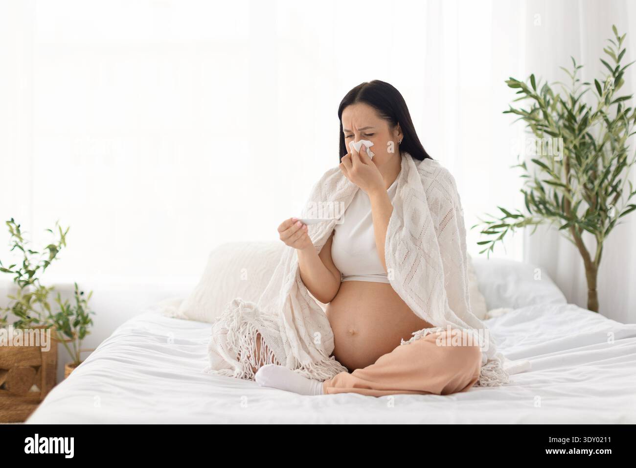 Schwangere Frau, die auf dem Bett sitzt, in eine Decke gehüllt und das Thermometer überprüft, sich unwohl fühlt Stockfoto