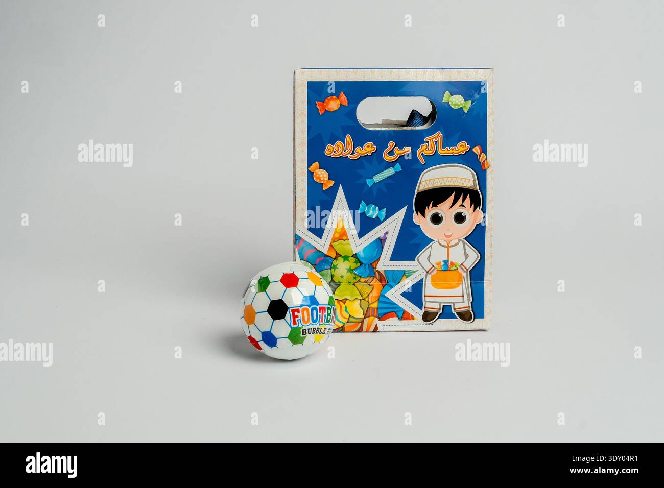 Fußball-Kugel-Luftpolsterspielzeug neben buntem Papppaket mit Zeichentrickjunge-Illustration und arabischem Text, isoliert auf grauem Hintergrund, Studio-Beleuchtung. Stockfoto