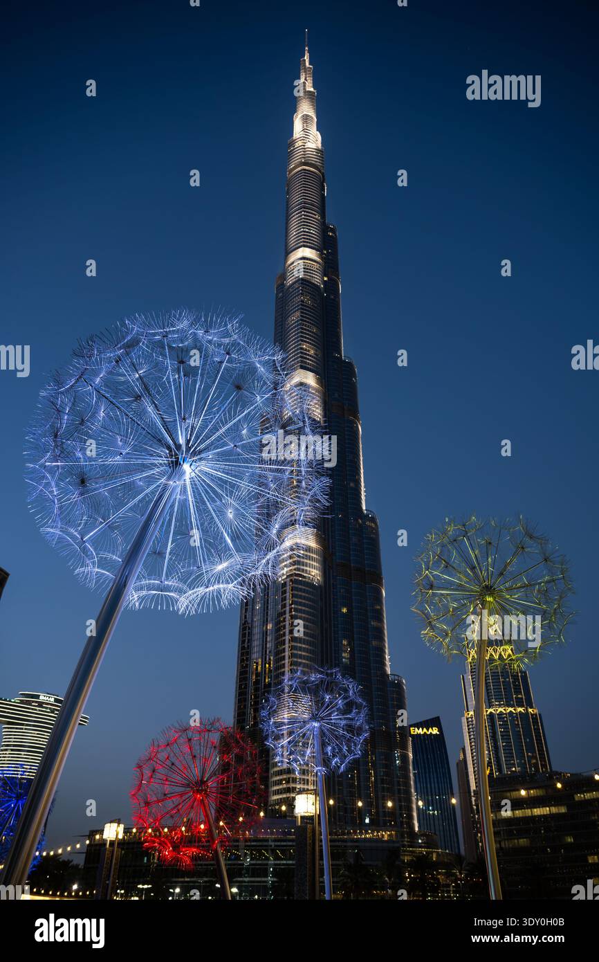 Ein niedriges, vertikales Foto des Burj Khalifa in Dubai bei Nacht, betrachtet durch große, beleuchtete Kunstinstallationen in Löwenzahn-Form. Das Licht Stockfoto