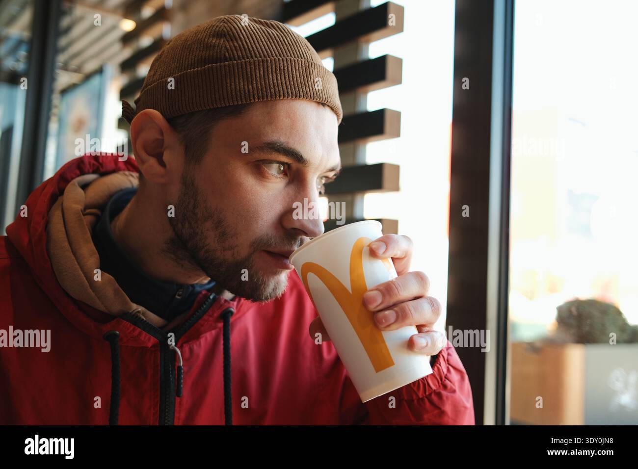 Ein Mann trinkt Limonade aus einem gebrandeten Papierbecher, während er in der Nähe des Fensters im McDonalds Restaurant sitzt. Konzept der globalen Fast-Food-Kette und der schnellen städtischen Mahlzeit Stockfoto