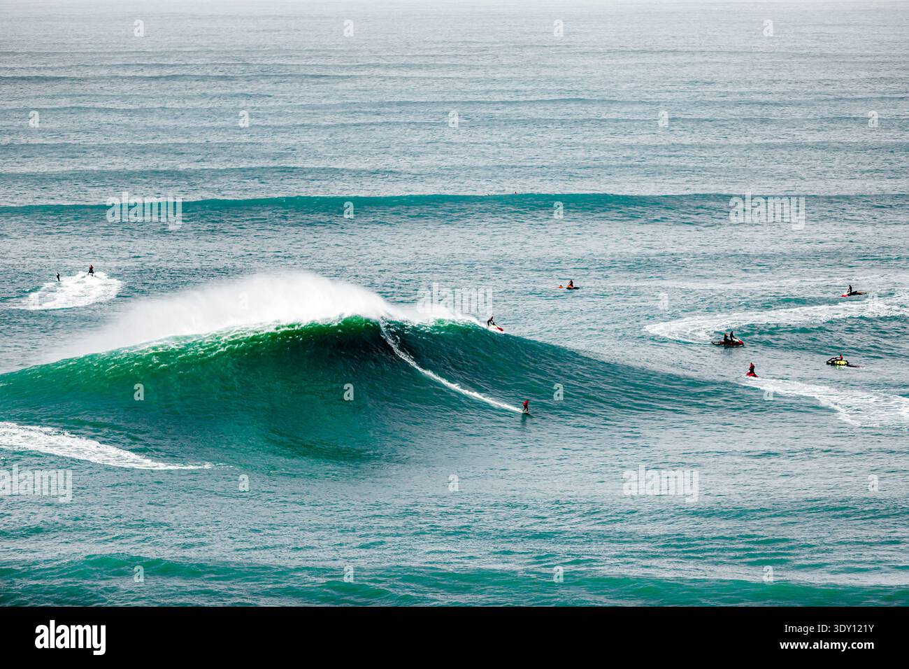 Surfer reiten türkisfarbene Wellen mit dynamischer Energie. Eine Welle bricht kraftvoll und sprüht weißen Schaum. Andere paddeln oder warten auf perfekte Bedingungen. Die s Stockfoto