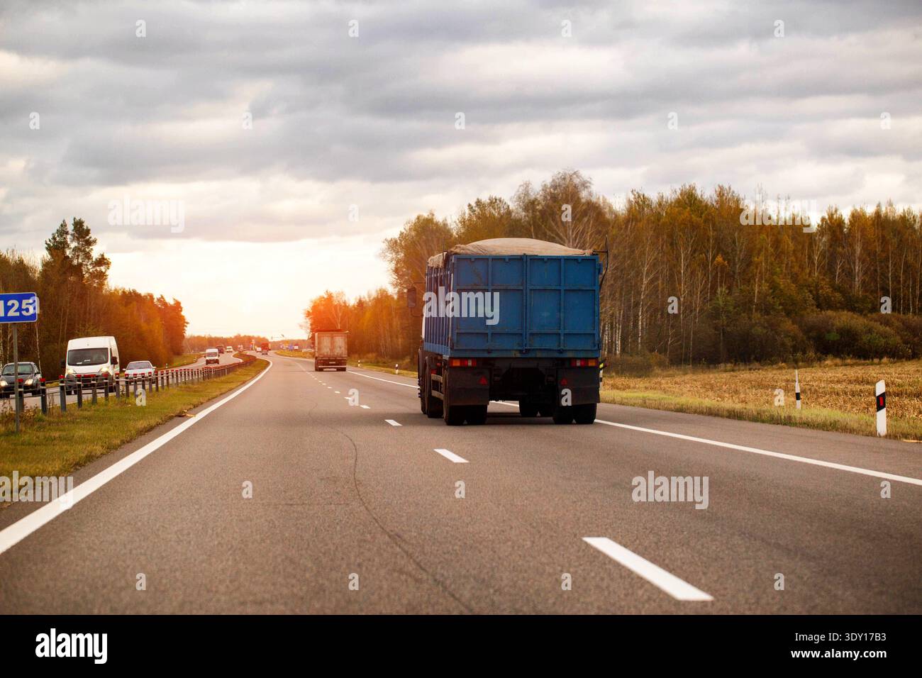 Rückansicht des blauen Muldenkippers, der Materialien auf Asphaltautobahn transportiert, umliegende Landschaft mit Bäumen mit farbenfrohem Herbstlaub, offenes Feld, andere Stockfoto