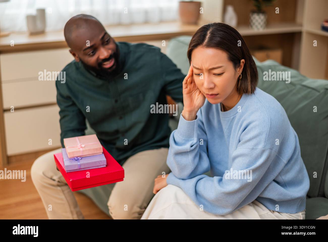 Eine enttäuschte Frau wendet sich auf dem Sofa ab, während ihr Partner einen Stapel Geschenkkartons anbietet, um einen angespannten Moment in ihrer Beziehung zu Hause festzuhalten. Stockfoto
