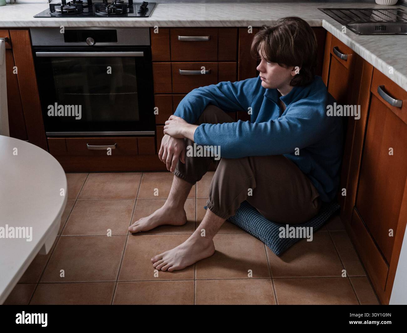 Junger Mann, der allein auf dem Küchenboden sitzt und wegschaut. Konzept von Depression, Einsamkeit, psychischen Gesundheitskämpfen und emotionalem Stress zu Hause. Stockfoto