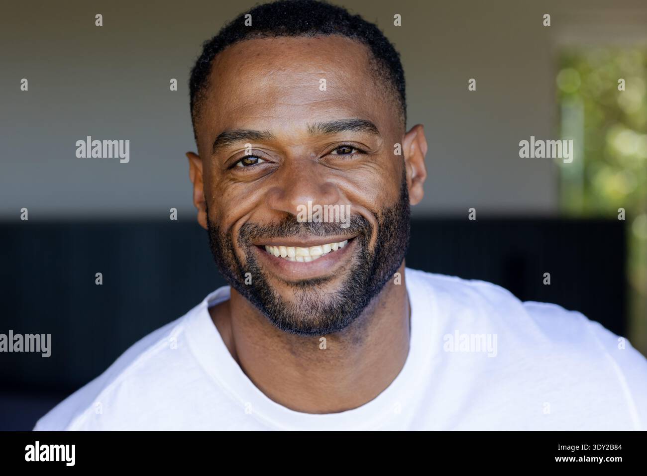 Afroamerikaner lächelt, während er ein weißes T-Shirt auf der schattigen Veranda trägt, Sonnenlicht filtert. Kopfschuss, bärtig, warm, natürlich, Outdoor, umweltfreundlicher Stockfoto