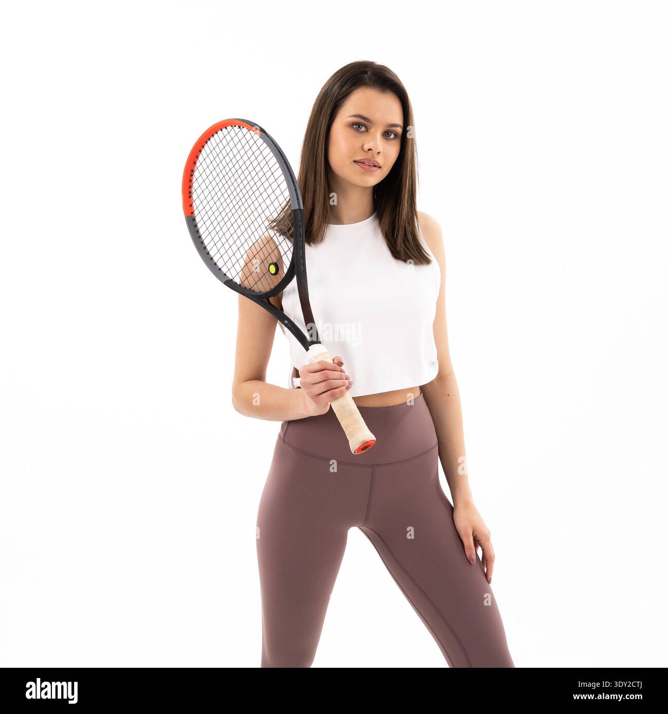 Die junge Frau hält Tennisschläger mit konzentriertem Ausdruck. Sie trägt passgenaue Sportausrüstung und steht selbstbewusst. Ein reiner weißer Hintergrund hebt sie hervor Stockfoto
