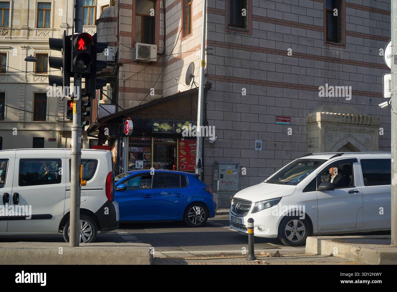 Rote Fußgängerampel auf der Mürselpaşa Ave, Fatih. Ein weißer Mercedes-Van und ein blaues Auto fahren an einem historischen Steingebäude in Istanbul vorbei. Stockfoto