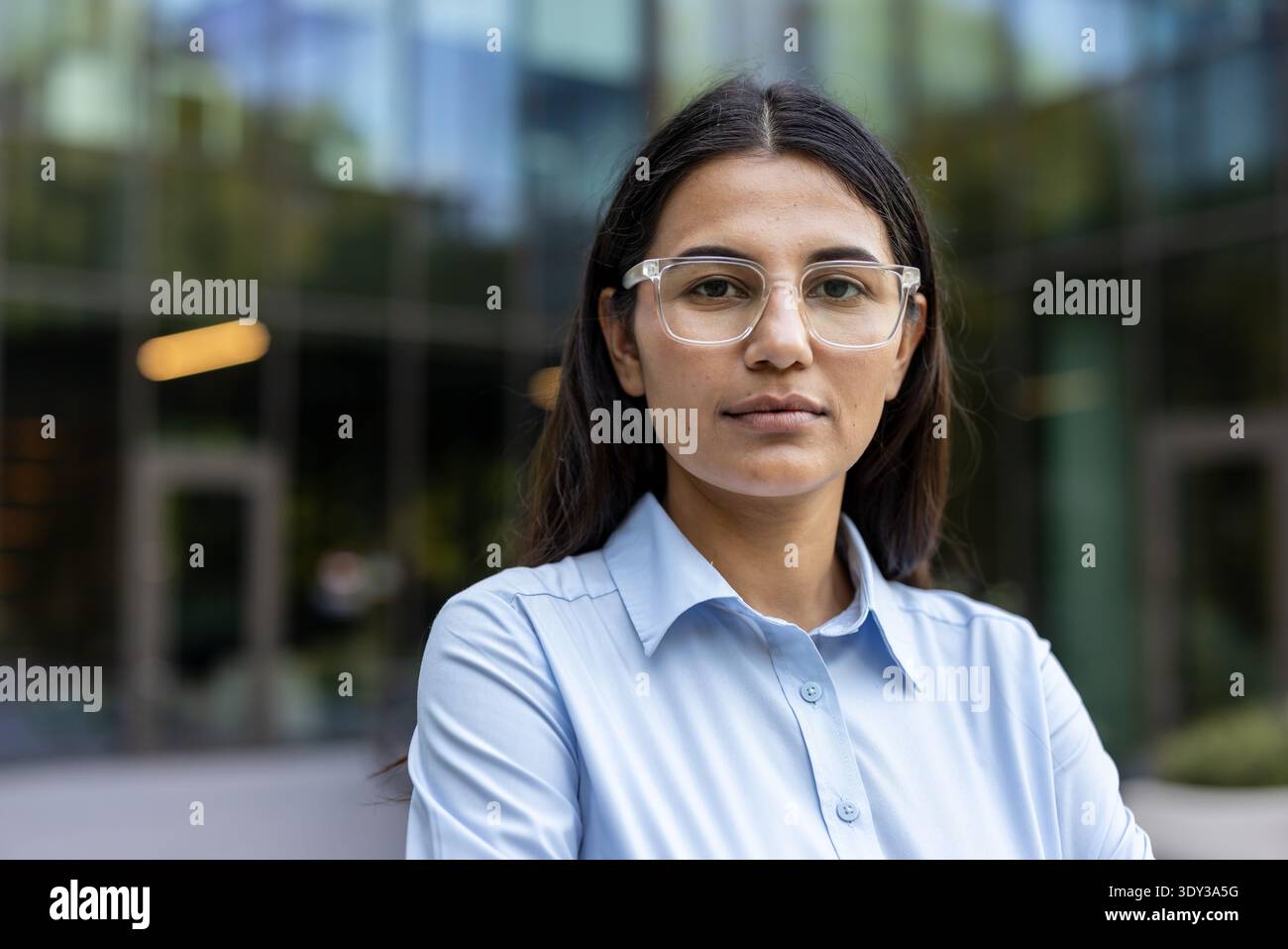Eine junge Frau, die ein hellblaues Hemd und eine klar gerahmte Brille trägt. Selbstbewusst im Freien vor einem modernen Glasbürogebäude stehen. Darstellung von Entschlossenheit und unternehmerischer Expertise Stockfoto