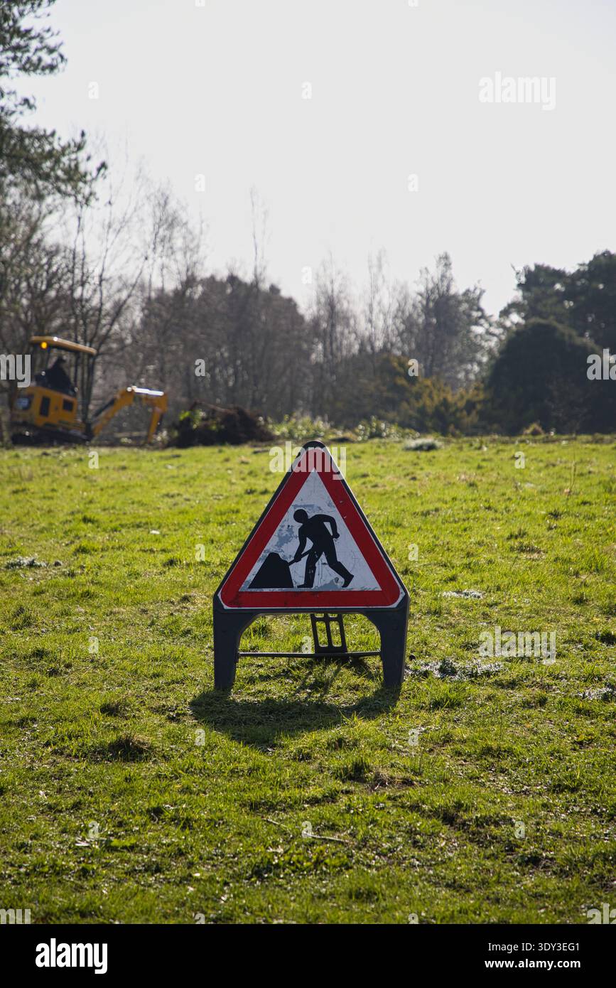 Ein Warnschild für Männer bei der Arbeit auf einem grasbewachsenen Feld in Rivington Terrace Gardens, Bolton, Großbritannien, mit einem gelben Minibagger im Hintergrund. Stockfoto