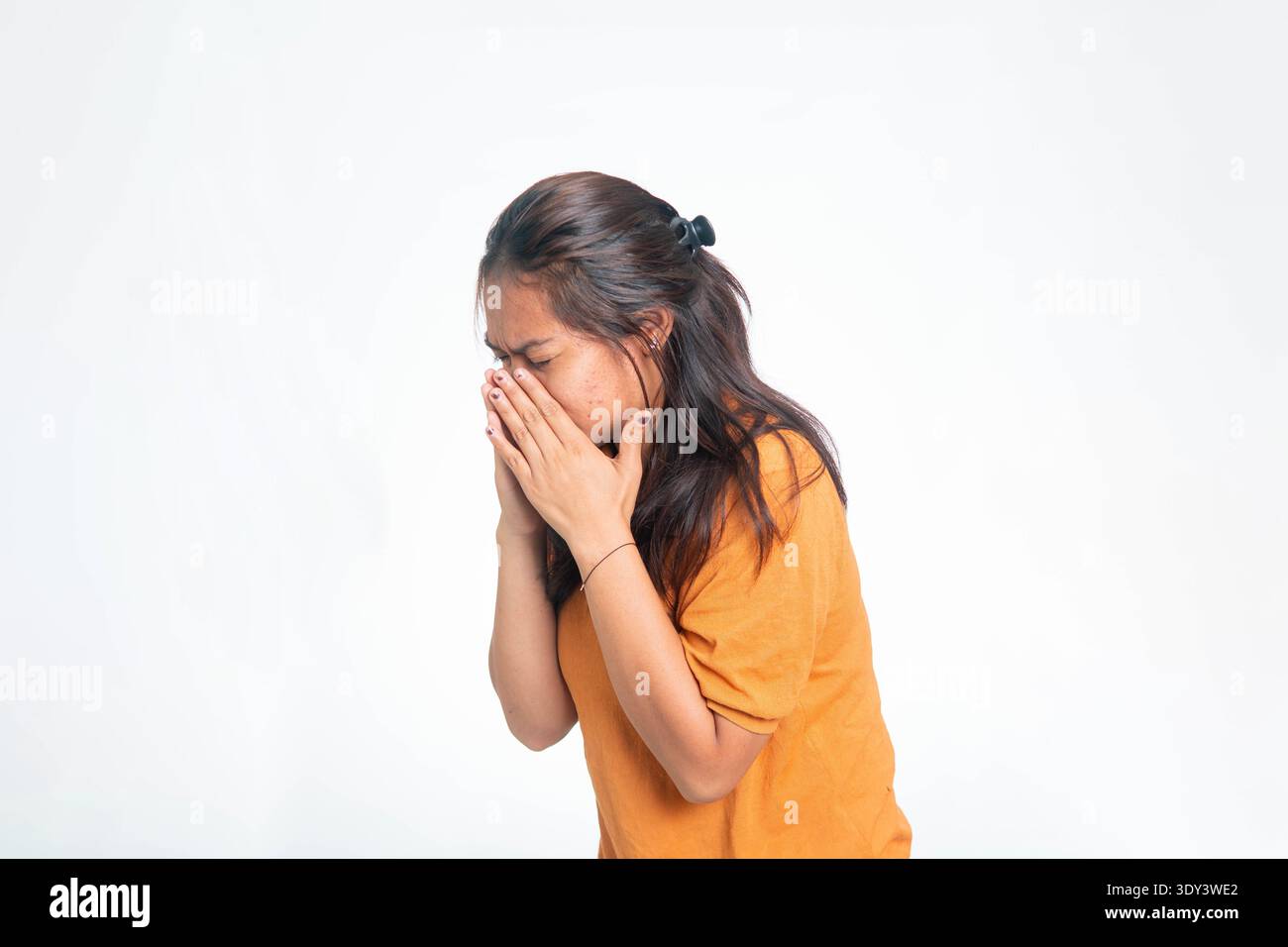 Junge indonesische Frau in orangefarbenem Hemd niesend und ihr Gesicht mit Händen bedeckend, an Allergie oder saisonaler Krankheit leidend. Stockfoto