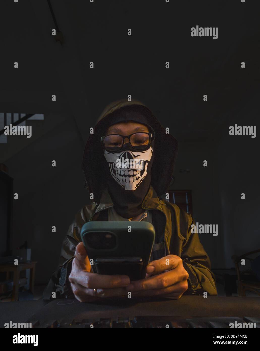 Eine Person, die eine Gesichtsmaske mit Schädelmuster, eine Brille und eine Kapuzenjacke trägt und aktiv mit einem leuchtenden Smartphone-Bildschirm interagiert. Die intensive Beleuchtung von t Stockfoto