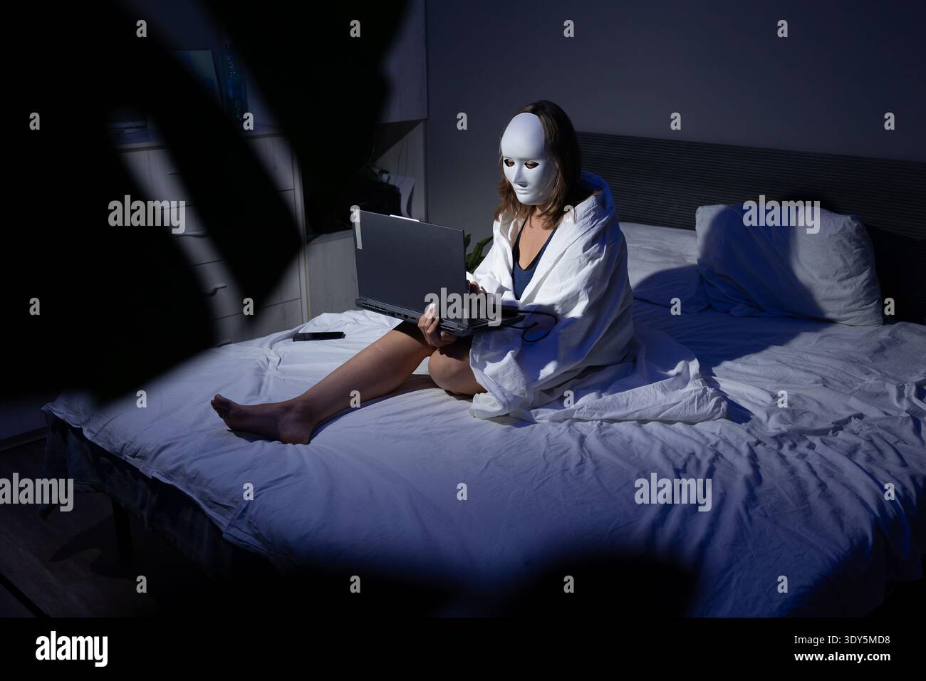 In einem schwach beleuchteten Schlafzimmer sitzt eine Person mit einer Maske auf einem Bett und konzentriert sich auf einen Laptop. Sie tragen einen Bademantel und stellen das wachsende Anliegen der digitalen Privatsphäre und des Datenschutzes dar. Stockfoto