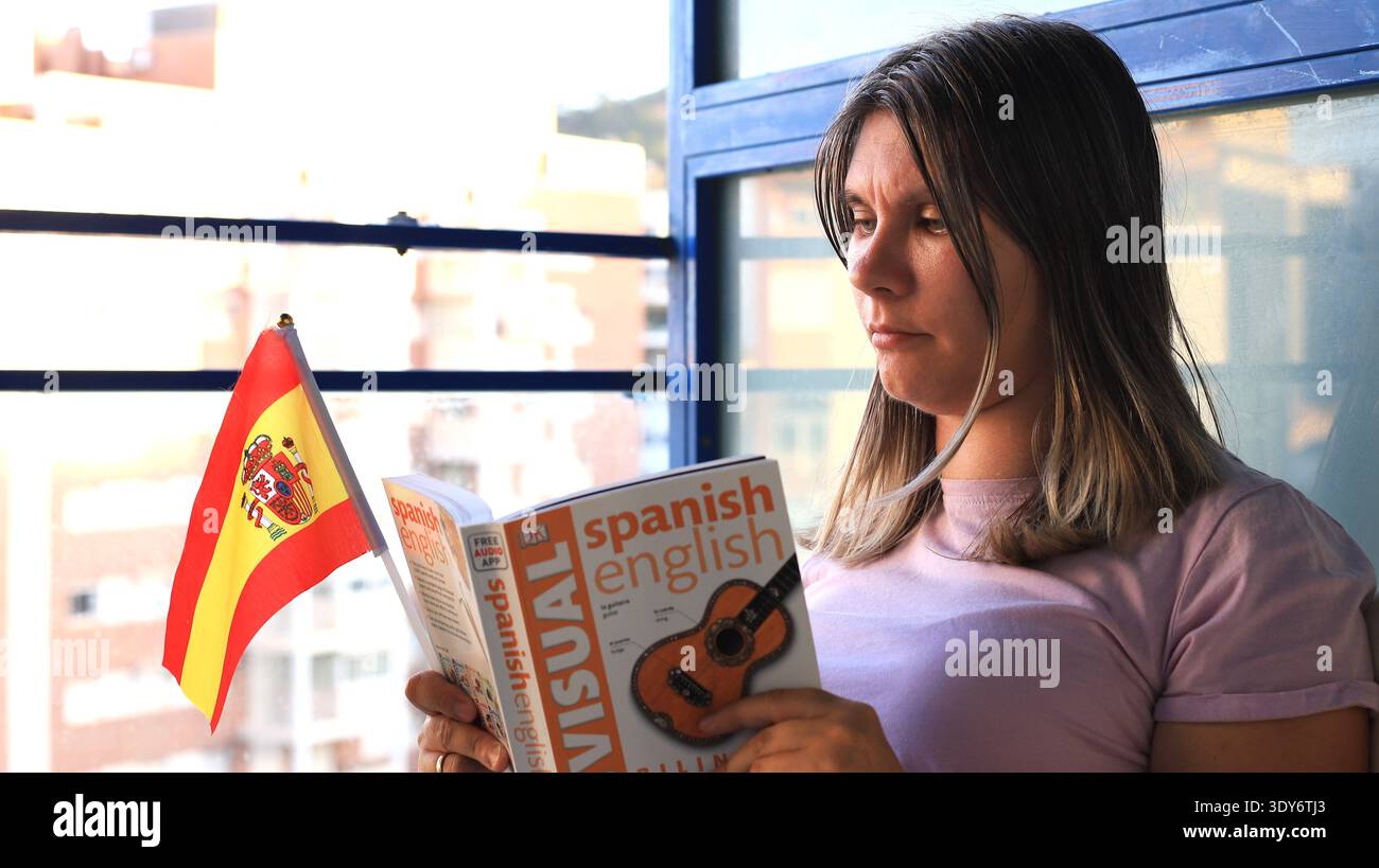 Junge Frau, die ein Spanisch-Englisch-Bildwörterbuch liest, während sie eine kleine spanische Flagge hält Stockfoto