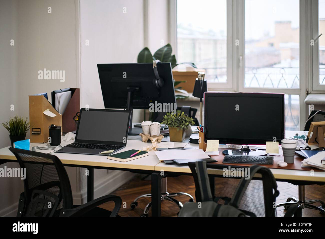 Ein leerer Büroarbeitsplatz zeigt zwei Schreibtische mit Computern, verstreuten Dokumenten, Büromaterialien, Topfpflanzen und Reinigungsmaterialien, was auf kürzlich durchgeführte oder laufende Reinigungsaktivitäten hindeutet Stockfoto