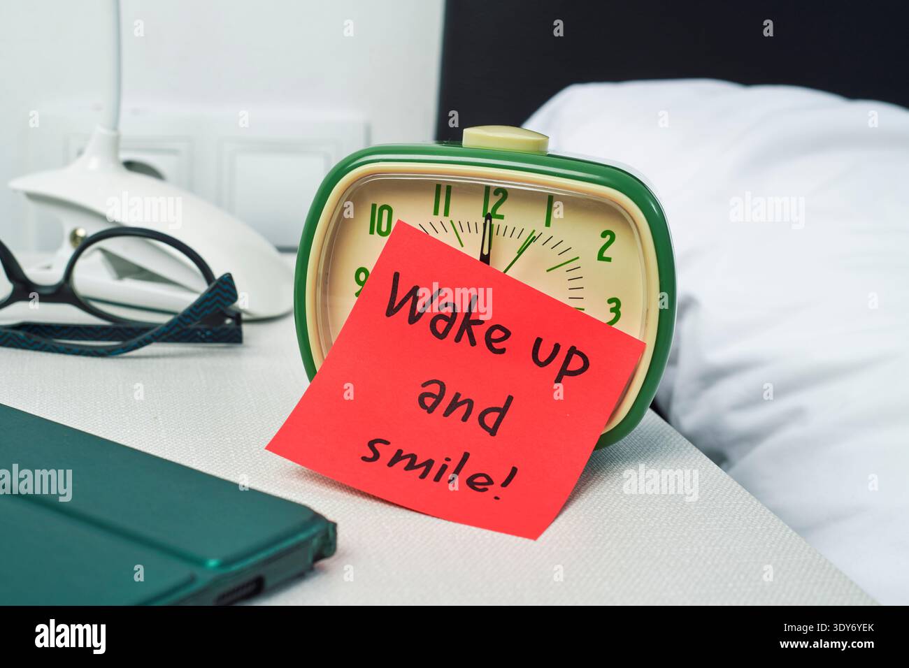 Eine rote Haftnotiz mit der Nachricht Wake up and Smile lehnt sich an einen grünen Wecker auf einem Nachttisch, in der Nähe befinden sich eine Brille und eine weiße Lampe Stockfoto