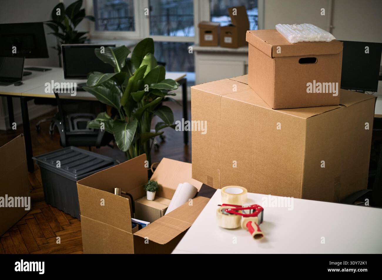 Gestapelte Kartonagen und Reinigungsmittel belegen moderne Bürobereiche, Verpackungsmaterial und Bandspender auf dem Schreibtisch, Innenanlage sichtbar unter beweglichen Gegenständen Stockfoto