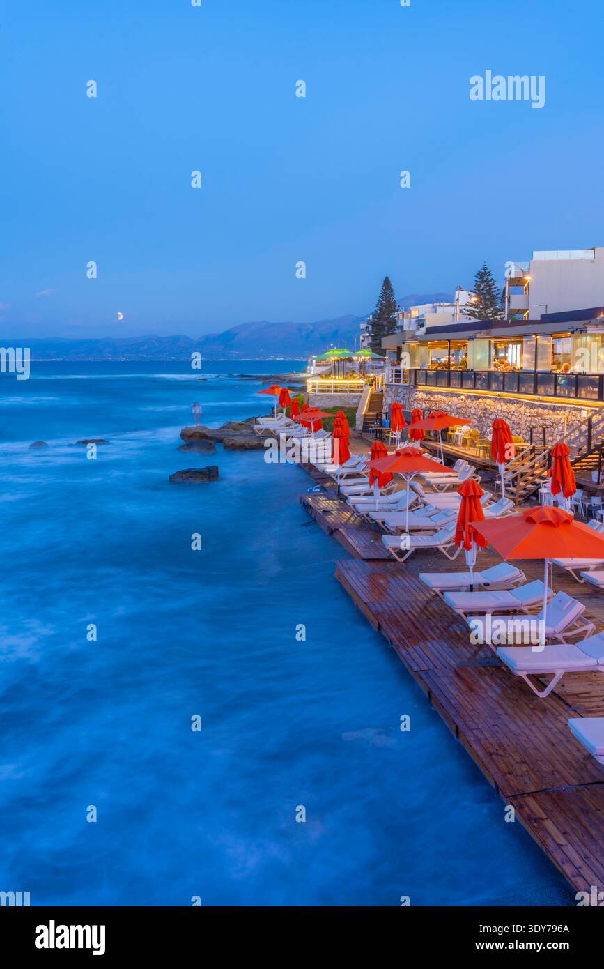 Taverne am Meer in der Abenddämmerung mit orangefarbenen Sonnenschirmen in Limenas Chersonisou, Kreta, Griechenland Stockfoto