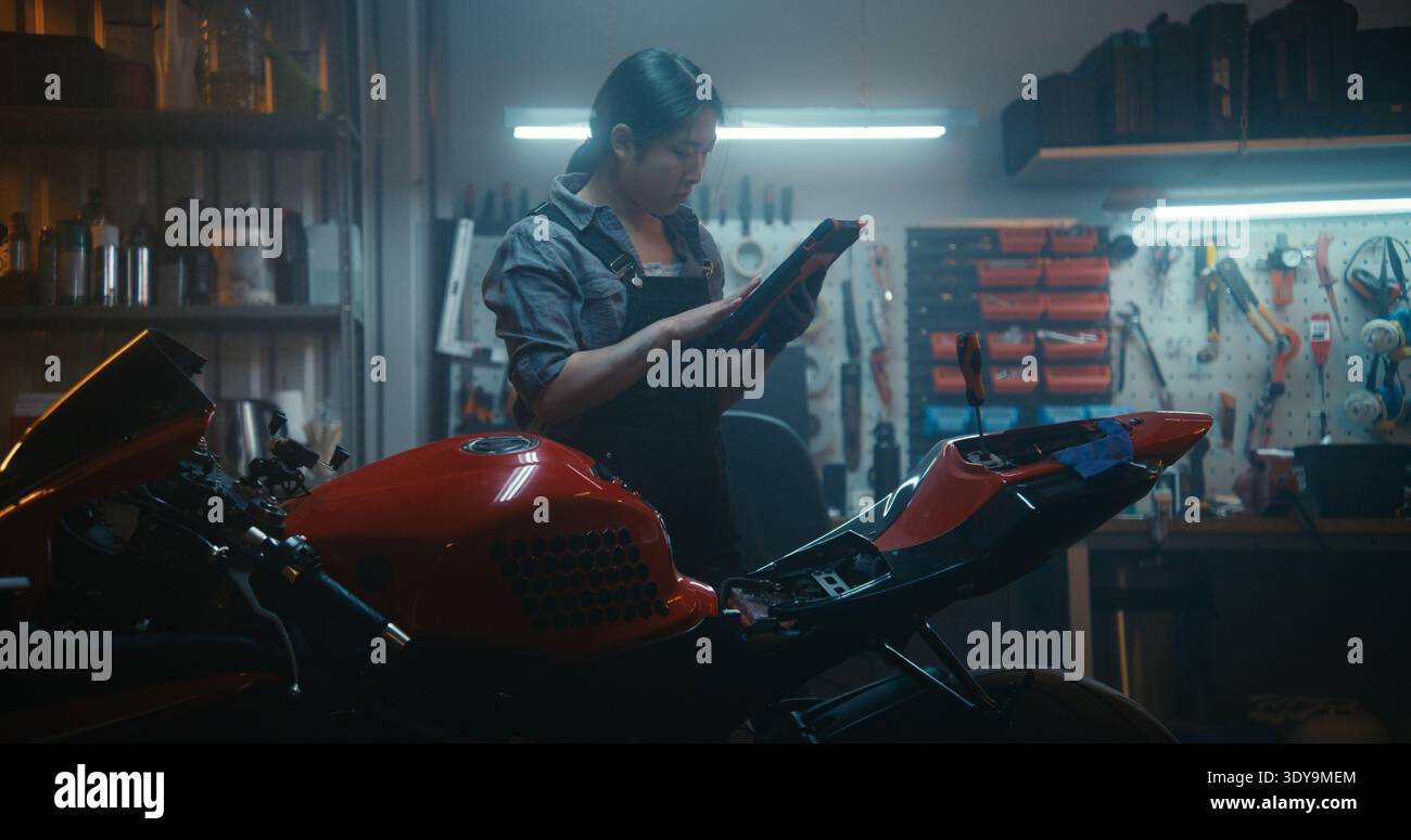 Asiatische Bikerin führt komplexe Aufgaben auf dem elektrischen System des Fahrrads durch und vergleicht die tatsächliche Batterieverkabelung mit 3D-Handbuch auf dem Tablet in der Garage mit gut ausgestatteten Werkzeugen. Professioneller Ansatz bei der Fahrzeugpflege. Stockfoto