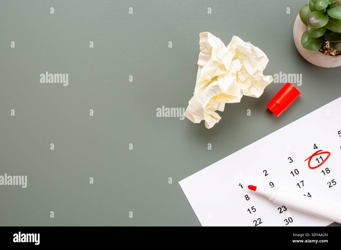 Hochwinkelansicht einer zerknitterten Papierkugel, eines roten Markers und eines Kalenders mit eingekreistem Datum auf einer olivgrünen Oberfläche mit Kopierraum. Stockfoto