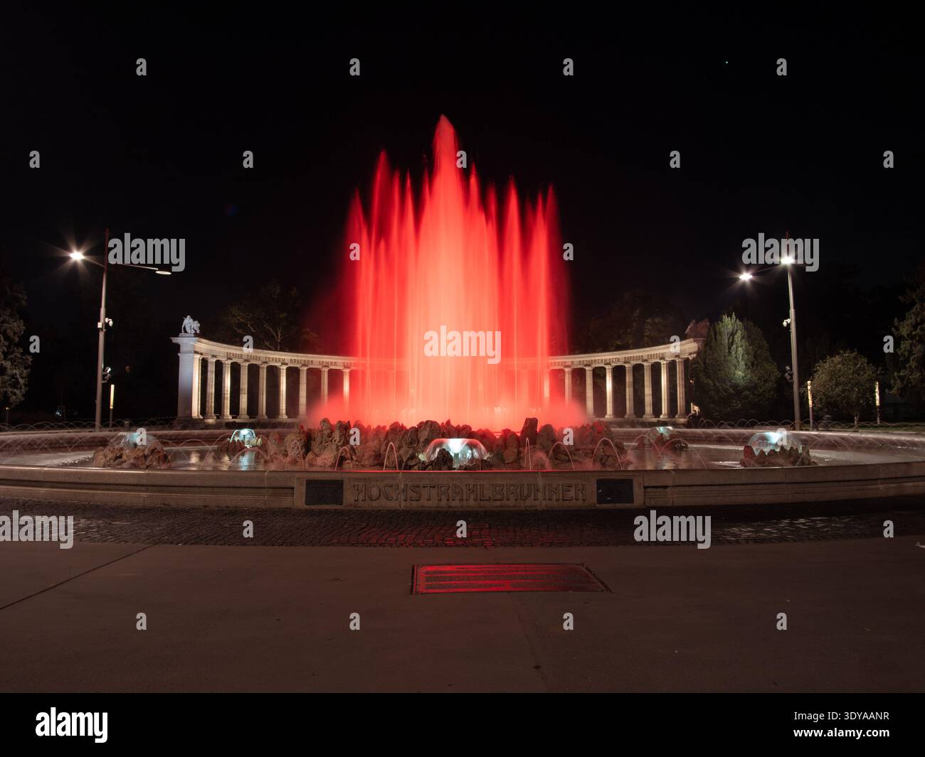 Hochstrahlbrunnen Wien bei Nacht: Beleuchtete rote Springbrunnenstrahlen, historische Kolonnade und Statuen, dramatische Rotlichtdarstellung. Stockfoto