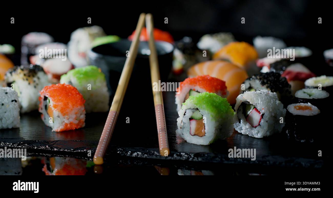 Farbenfrohe Auswahl an Sushi, serviert auf Schieferplatte mit Essstäbchen, die in einem Speisesaal während der Abendstunden daneben stehen Stockfoto