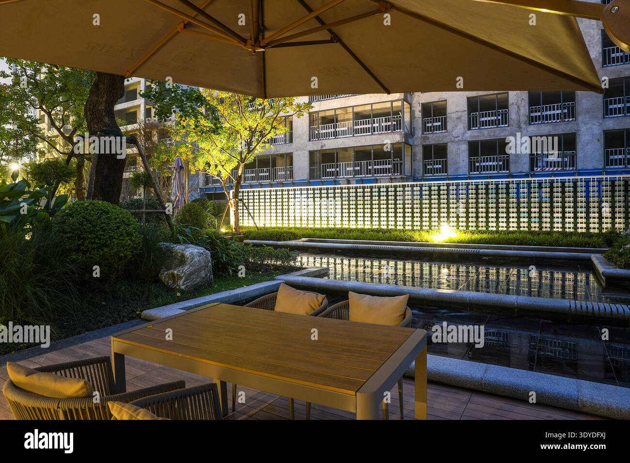 Eine intime Atmosphäre auf einer Terrasse mit Blick auf einen reflektierenden Pool und eine leuchtende architektonische Wand. Das Bild vermittelt Raffinesse, Luxus, Stockfoto