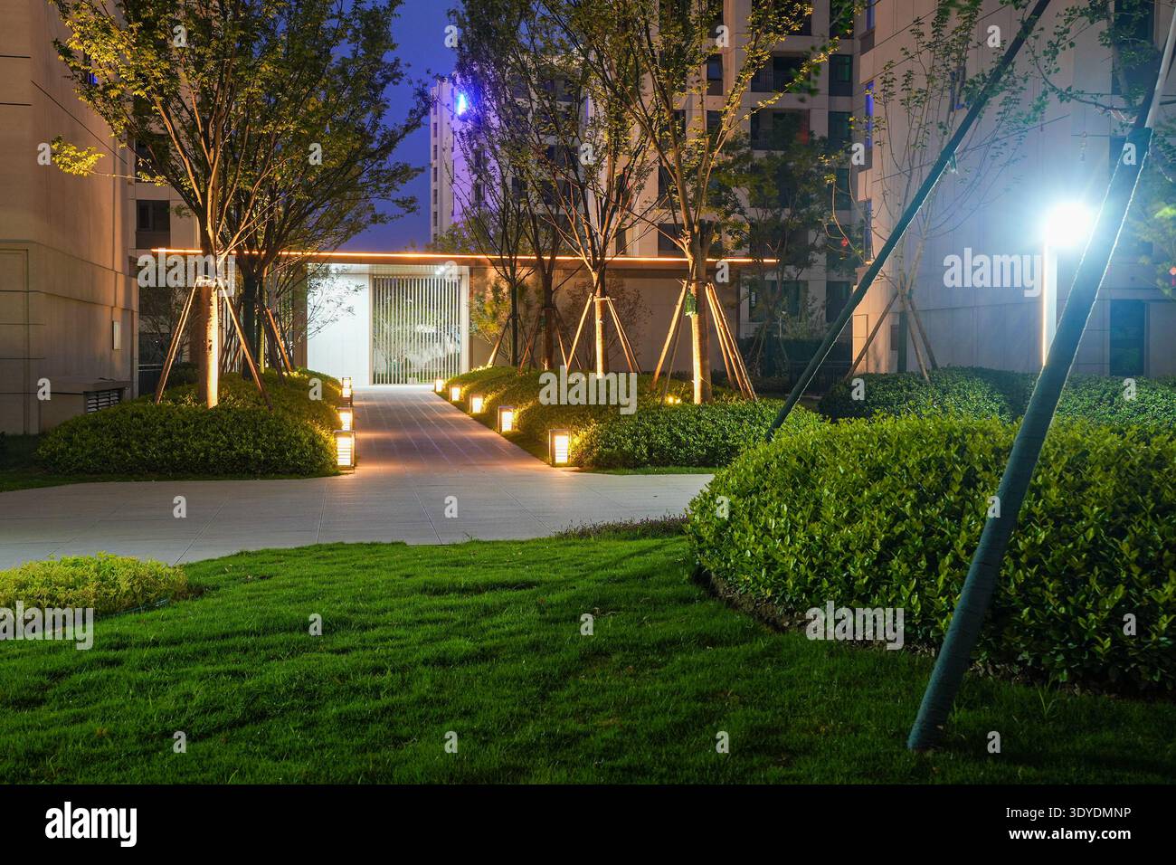 Ein einladender Eingang zu einem Wohnpark mit warmer Beleuchtung an Bäumen und Wegen am Abend. Das Bild vermittelt Gastfreundschaft, Gemeinschaftssicherheit Stockfoto