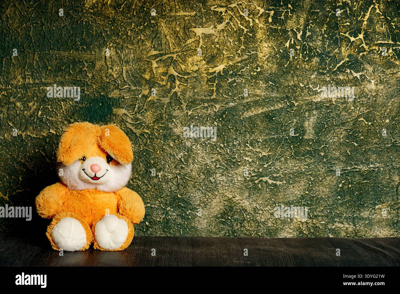 Ein weicher, oranger gefüllter Häschen sitzt auf einer dunklen Oberfläche vor einer grünen Wand mit rauer Textur. Stockfoto