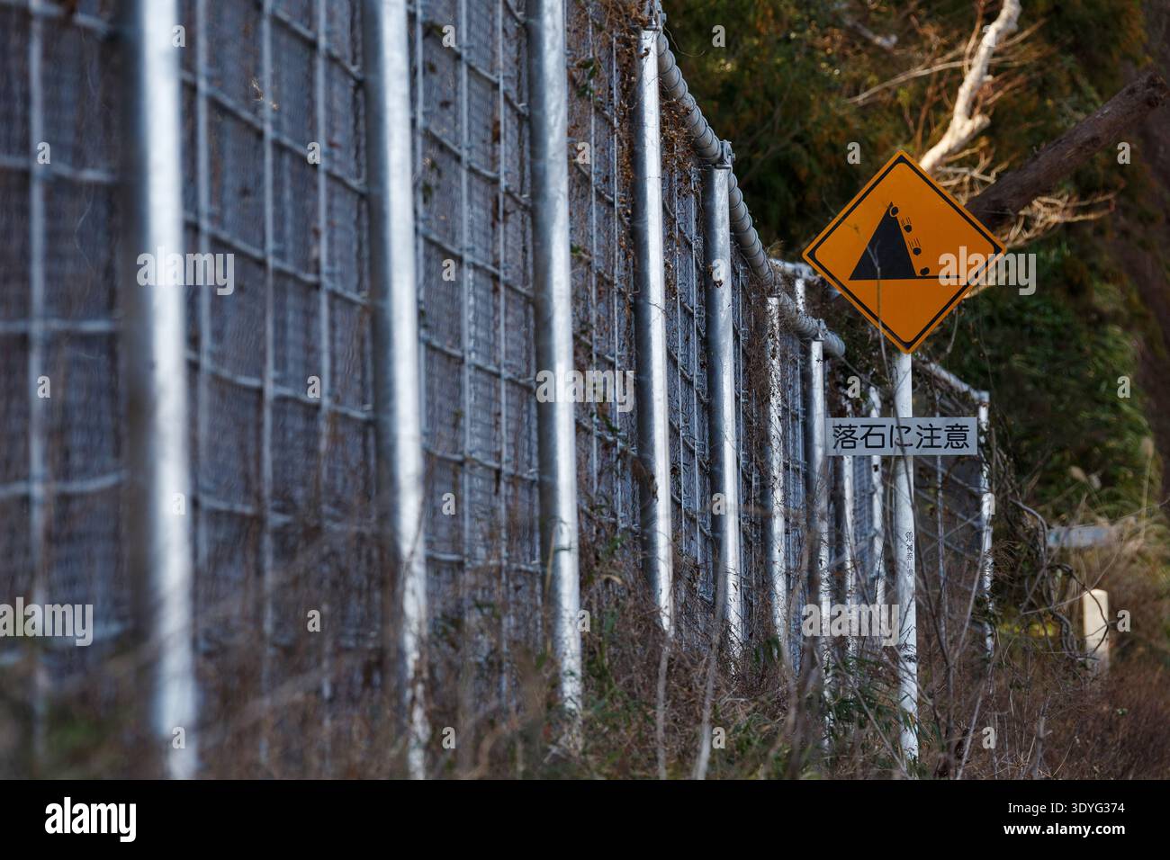 Ein großer Metallzaun zum Fangen herabfallender Felsen durch ein Schild, das vor herabfallenden Felsen warnt, am Straßenrand in der Nähe von Aikara im ländlichen Kanagawa, Japan. Stockfoto