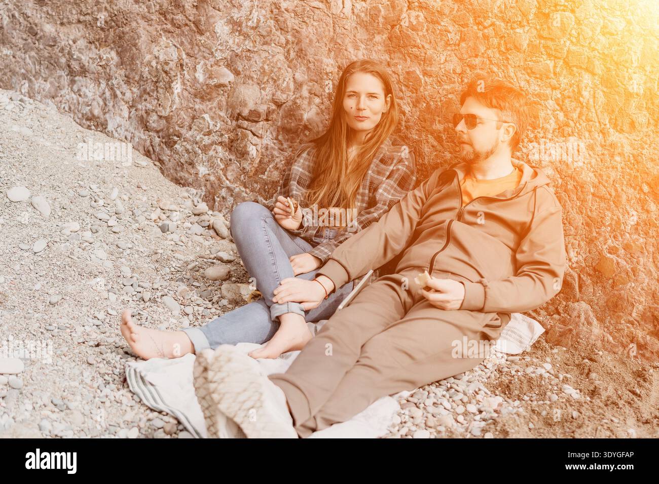 Paare Strandentspannung junger Mann und Frau, die am Kiesstrand vor felsigen Klippen sitzen und Snacks im goldenen Sonnenlicht teilen Stockfoto