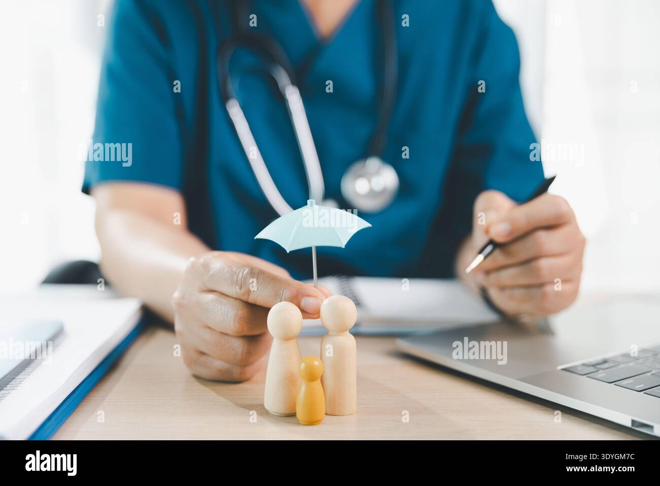 Medizinisches Personal schützt Holzfiguren mit Minischirm am Kliniktisch mit Stethoskop und Laptop, symbolisiert Krankenversicherung, Risk cov Stockfoto