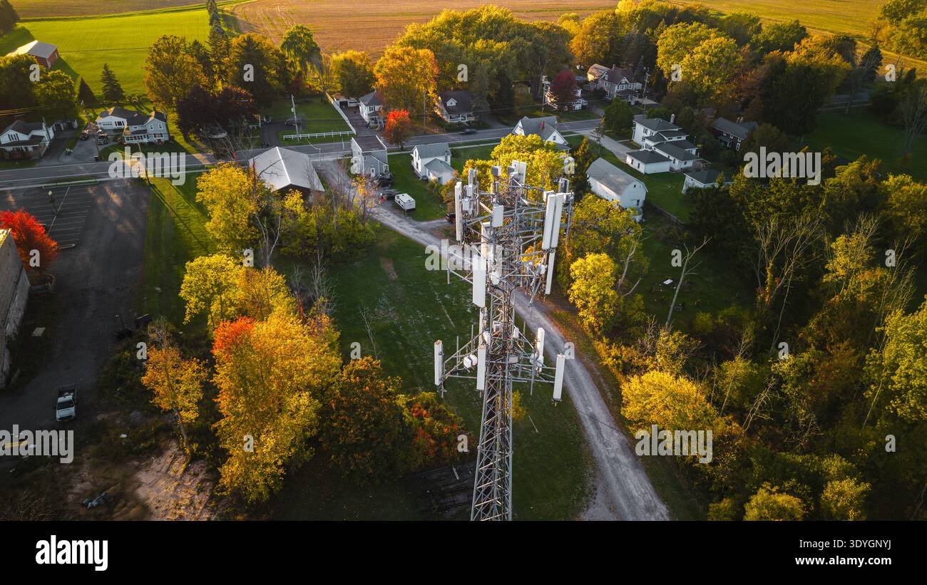 Kommunikationsturm in der Nähe kleiner Vorstadthäuser, umgeben von Herbstbäumen und grünen Feldern, von oben im warmen Abendlicht erfasst. Stockfoto
