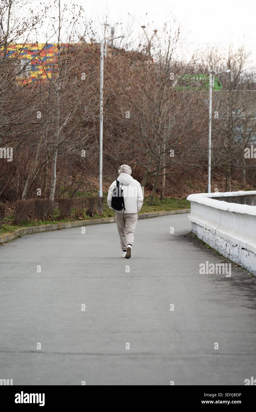 Rückansicht eines jungen Menschen mit weißem Hoodie und Kopfhörern, der an einem bewölkten Tag allein in einem Stadtpark spaziert. Moderner urbaner Lebensstil: Ein Mann mit Draht Stockfoto