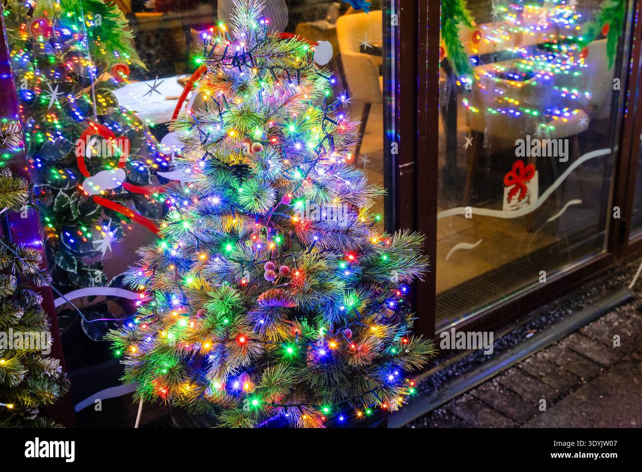 Farbenfroher Weihnachtsbaum mit elektrischen Girlanden auf der Stadtstraße in der Nähe von Schaufenstern in der Winternacht Stockfoto