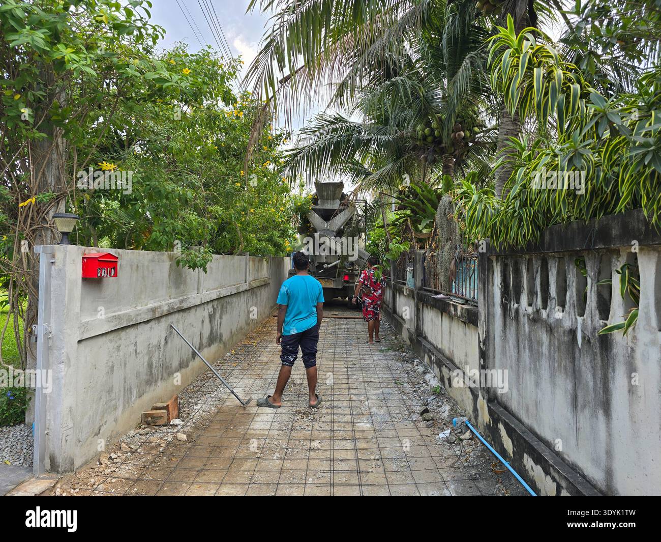 Ein Arbeiter gießt Beton auf einen neu angelegten Weg, der von üppiger Vegetation in Bang Lamung umgeben ist. Die tropische Atmosphäre zeigt Palmen und farbenfrohe Pflanzen unter einem strahlenden Himmel. Stockfoto