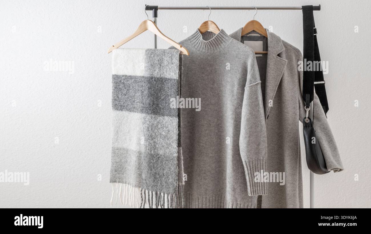 Hellgrauer Wollmantel oder -Jacke, Merino-Pullover oder -Kleid, Alpaka-Schal und schwarze Ledertasche auf Kleiderbügel vor weißem Hintergrund. Herbst und Winter Stockfoto
