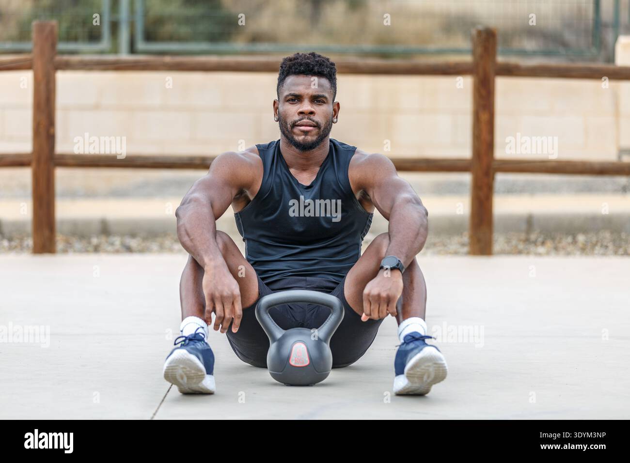 Voller Körper eines jungen bärtigen muskulösen afroamerikanischen Athleten in Sportbekleidung, der mit einer Kettlebell auf dem Boden sitzt und in die Kamera blickt Stockfoto