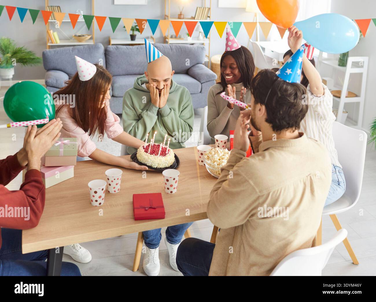 Aufgeregter Mann, der sein Gesicht mit Händen bedeckt, während Freunde in Partyhüten ihn mit Geburtstagskuchen überraschen. Stockfoto