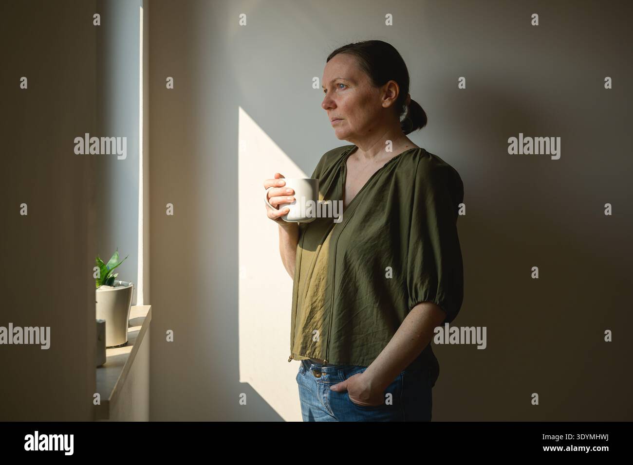 Frau mittleren Alters mit Pferdeschwanz, die Keramikbecher in der Nähe des Fensters hält, goldenes Morgenlicht und ruhige, introspektive Stimmung in friedlicher Inneneinrichtung. Selektiv Stockfoto